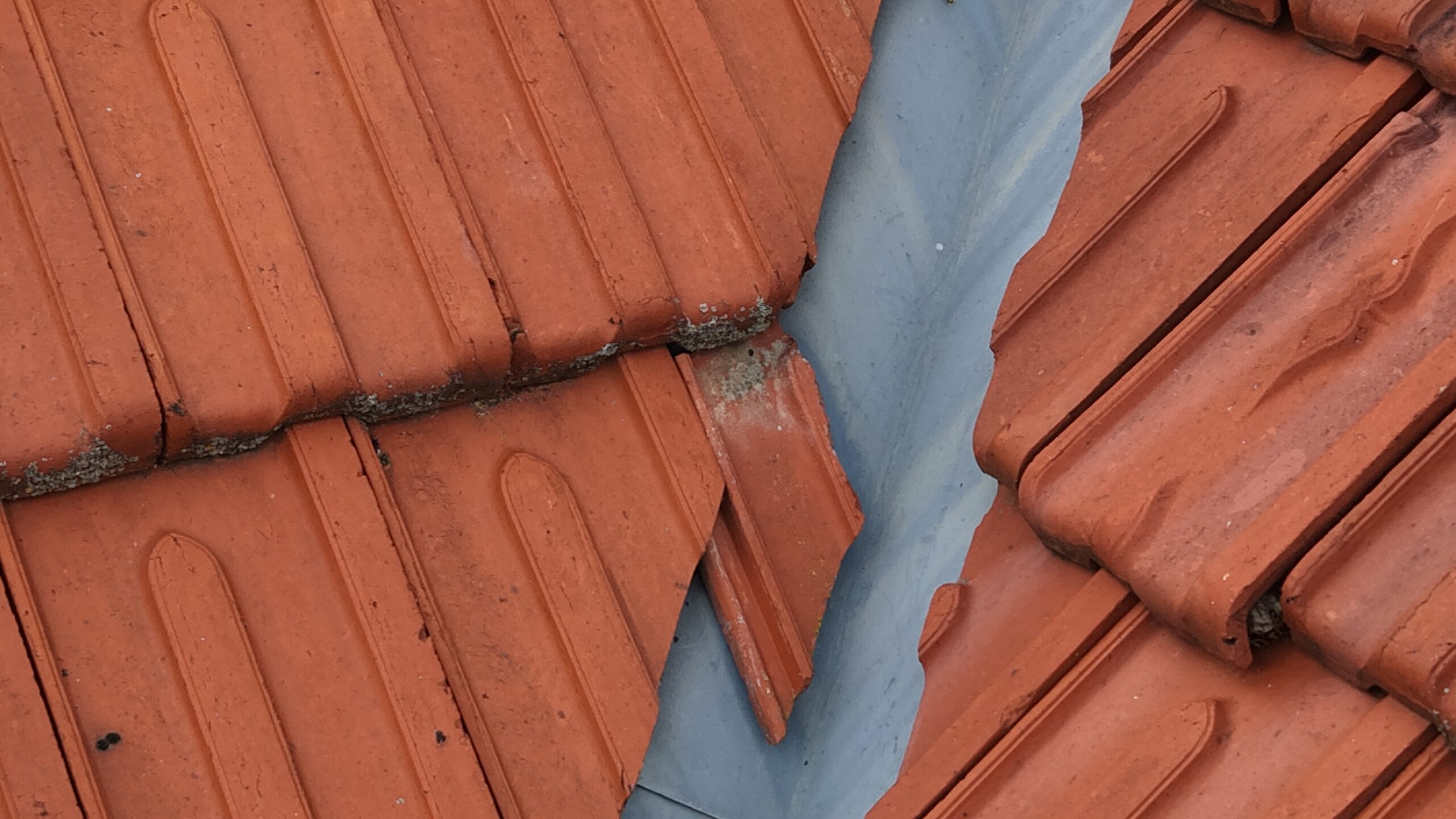 Vue rapprochée d’une toiture en tuiles montrant une jonction avec une bande d’étanchéité en zinc et des traces de dégradation visibles.
